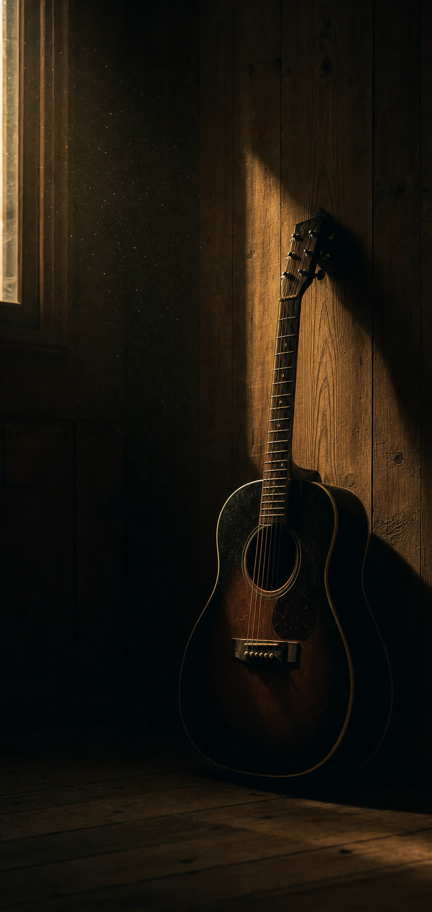 An acoustic guitar leaning against a wood-panelled wall in a dim room, lit by a narrow shaft of daylight.